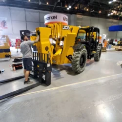 A man in a gray shirt is attaching a forklift attachment to a yellow JCB telehandler inside a large industrial building.
